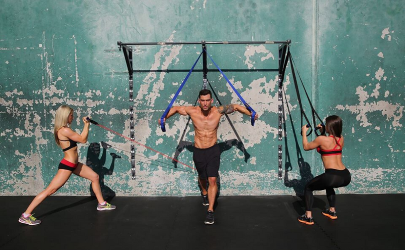 Attractive guy working out with bands attached to pull up bars. On both sides of him are two attractive women who are working out with bands attached to a concrete wall